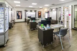A room with wooden floors and furniture, including desks, chairs, and a cabinet, is displayed with a monitor, a box, and other items. There are also posters on the wall, a cabinet with many eyeglasses, and a mirror.