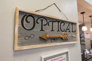 An optician shop sign hanging on a wall with a gray background, black text, and a wooden arrow with eyeglasses on either side.