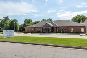 An exterior view of the Laster Eye Center in Oklahoma City, Oklahoma, with a sign, lush green lawn, brick walls, and trees in the distance under a blue sky with clouds.