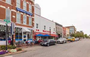 A busy street with cars parked on the side and people sitting outside a cafe and a shop on Main Street, the cafe has a blue sign and a blue umbrella while the shop has a red awning with the text 5-10 Waltons F&D
