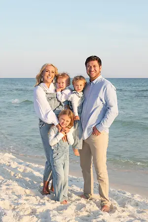 A family of four is standing on the beach, with the father holding a baby on his shoulders, the mother holding a toddler, and the other child standing beside them. They are all smiling and looking at the camera.