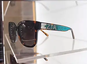 Black sunglasses with turquoise decorative detailing displayed on a clear acrylic stand