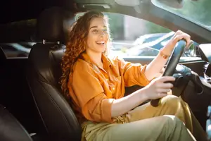 A woman is sitting inside a car and smiling while holding the steering wheel