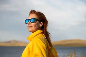 Woman in a yellow jacket standing near a lake with blue sunglasses