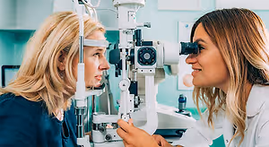 A woman in a white coat is examining another woman's eyes with a slit lamp in an eye clinic.