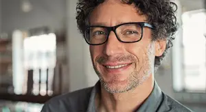 Man with curly hair wearing glasses and smiling in front of a white wall