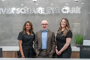 Three people standing next to each other in front of a sign that reads Never Square Eye Care