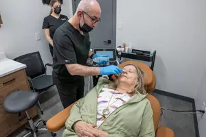 A man wearing a mask and gloves is examining a woman's eye while she is sitting in a chair in a room.