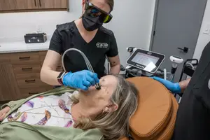 A woman is sitting in a chair with a medical professional wearing a mask and gloves, using a device to examine her face.