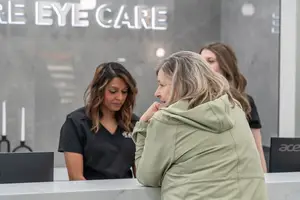 Three women are standing near a reception desk in an eye care clinic, with one woman looking towards the camera while another stands behind the desk with a laptop and two candles.