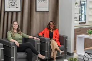 Two women in a room are sitting on a couch, one is wearing glasses and smiling, while the other is wearing a blazer and looking at the camera, with picture frames and a table with a potted plant behind them.