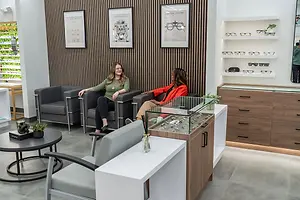 Two women sitting on chairs in a room with gray walls, wooden cabinets, and a table with a potted plant, and a shelf with eyeglasses in a retail shop.