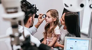 An ophthalmologist is examining a young girl's eyes with a slit lamp while her mother watches closely in a clinical setting.