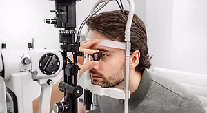 A man in a gray shirt looks into an ophthalmoscope while in a room with white walls.