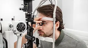 A man in a gray shirt looks into an ophthalmoscope while in a room with white walls.