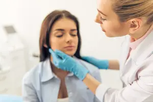 A woman wearing a white lab coat and blue gloves is examining the neck of another woman in a clinic.