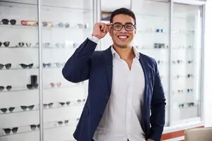 A smiling man wearing a navy blazer and white shirt stands in front of a glass display case filled with sunglasses
