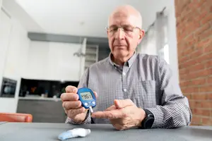 An elderly man is measuring his blood sugar using a digital glucometer in his kitchen.
