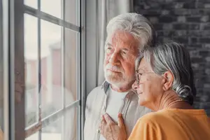 An elderly man and woman standing close together, looking out of a window and gazing at each other