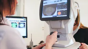 A woman in a white lab coat is adjusting a white machine with a screen, a woman in a black shirt is sitting in front of her, and a monitor is on the left with a ring-shaped display.