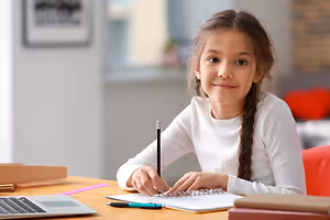 A girl sitting on a chair in front of a desk with a laptop and books, smiling while holding a pen and writing on a notebook