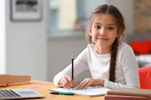 A girl sitting on a chair in front of a desk with a laptop and books, smiling while holding a pen and writing on a notebook