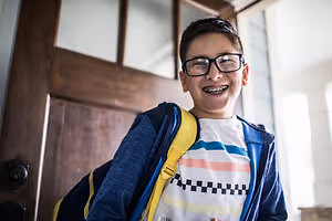 A boy with braces smiles and stands in front of a wooden door, wearing a backpack and glasses.
