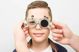 A young boy wearing green-framed glasses is having his vision tested by an adult in a white room.