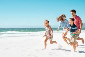 A family of four is running on the beach, the woman in the front is looking back and smiling, the man beside her is laughing, and the two kids are running ahead.
