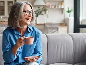 A smiling older woman with gray hair holding a pink coffee cup on a couch in a kitchen