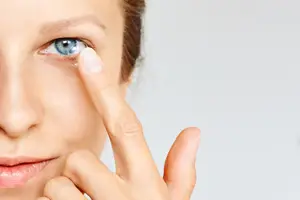 A woman is adjusting a contact lens in her left eye with her right hand. The woman is looking straight ahead, and her hair is tied back. The background is white, and she is wearing a red lip color.