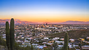Sunset over a city with a mountain range in the background, featuring a variety of buildings and cacti.