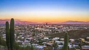 Sunset over a city with a mountain range in the background, featuring a variety of buildings and cacti.