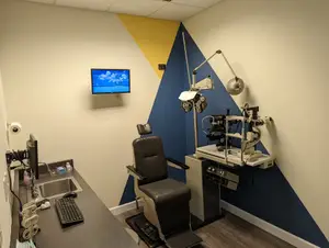 A medical examination room with a desk, computer, sink, chair, and eye examination machine on the wall.