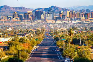 A busy city street with many buildings and cars driving by, surrounded by trees and mountains in the distance.