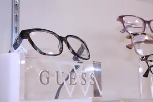 A pair of Guess eyeglasses are displayed on a clear stand with a price tag in a retail store.