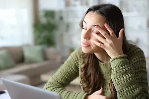 A woman sitting in front of a laptop with her eyes closed and her hands on her forehead.