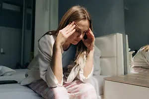 A woman sitting on a bed holding her head and looking upset