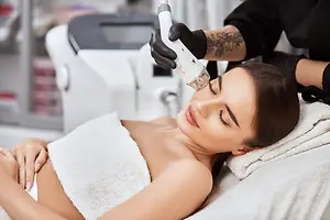 A woman lies on a bed in a salon, getting a facial treatment with a laser device.