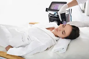 A woman is lying down on a bed wearing a white robe and receiving an eyebrow laser treatment