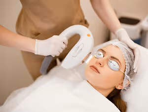 A woman is lying on a bed wearing a hairnet and goggles while a person is holding a white laser machine in their hands and looking at her.
