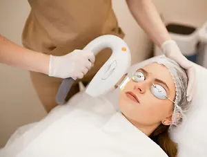 A woman is lying on a bed wearing a hairnet and goggles while a person is holding a white laser machine in their hands and looking at her.
