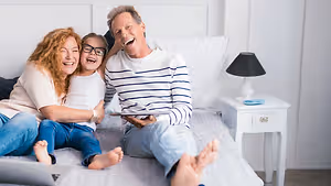 A family of three sitting on a bed, laughing and smiling while holding a tablet. A nightstand with a lamp and a plate is next to them. The girl in glasses is sitting on the father's lap. The bed is covered with a white sheet. The room has white walls, and the floor is gray.