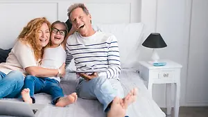 A family of three sitting on a bed, laughing and smiling while holding a tablet. A nightstand with a lamp and a plate is next to them. The girl in glasses is sitting on the father's lap. The bed is covered with a white sheet. The room has white walls, and the floor is gray.