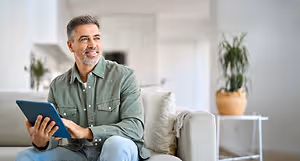 An adult male is sitting on a couch in his living room and holding a tablet with both hands, and there is a potted plant beside him on the right.