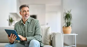 An adult male is sitting on a couch in his living room and holding a tablet with both hands, and there is a potted plant beside him on the right.