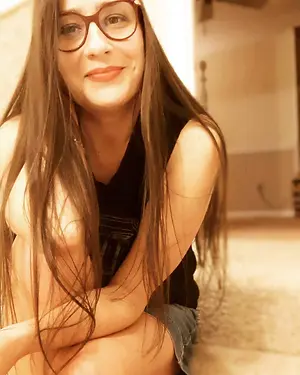 A woman with long hair wearing glasses and a black sleeveless top is sitting on a carpeted floor with her legs crossed, smiling and posing for a photo in a room with a white wall and a bed in the background.
