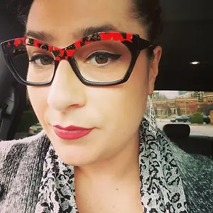Close-up of a woman with red lips, wearing black and white patterned glasses and scarf, and sitting inside a car