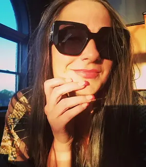A woman wearing sunglasses and a patterned shirt is posing for a photo with her hand on her face.