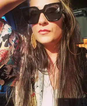 A woman with long hair is wearing sunglasses and earrings and is looking to the side. She is wearing a white top with a colorful pattern. Behind her is another person wearing a floral shirt and a necklace. The person's hand is touching the woman's head. The background is blurry, and it seems like they are in a room with a blue wall. The light is shining on the woman's face.